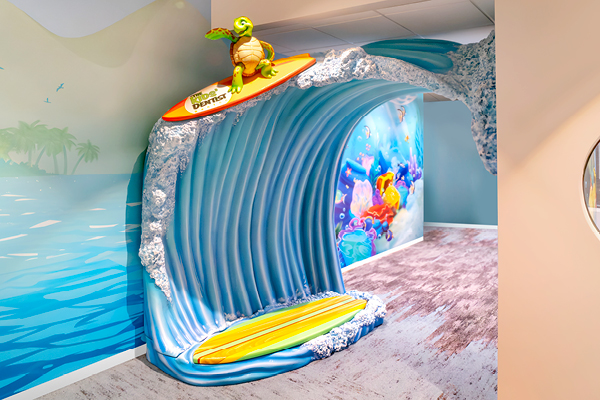 A playful indoor decor features a large blue wave with foam details, a turtle character on a surfboard, and ocean-themed wall art in the background, creating a fun, aquatic atmosphere.