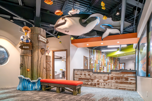 Childrens waiting area with a red bench, playful pool-themed decor, a large orca model hanging from the ceiling, and a wooden reception desk in the background. Bright, colorful, and inviting space.