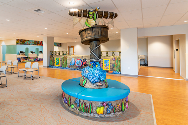 A colorful childrens waiting area features a circular blue bench with an ocean-themed sculpture and a turtle on top. The room has bright decor, chairs, and a reception desk in the background.