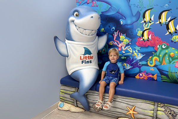 A young child sits on a blue bench next to a large friendly shark mascot statue wearing a Little Fins shirt, with an underwater-themed mural in the background.