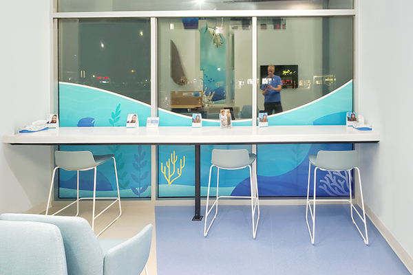 A modern, clean waiting area with a high counter, four light blue chairs, and ocean-themed wall art. A large window shows a nighttime view and a person reflected in the glass.