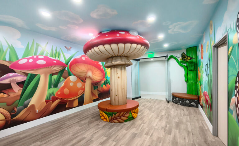A whimsical room with large, colorful mushroom decorations, a wooden mushroom-shaped seat, mural walls featuring grass, flowers, butterflies, and a tree door—perfect for an imaginative undersea kids ministry—complete with light wood flooring and bright lighting.
