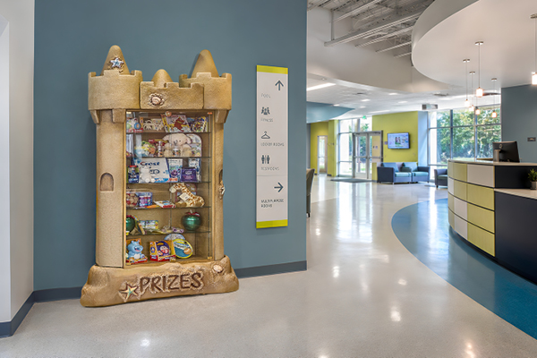 A sandcastle-shaped prize display case filled with toys and stuffed animals stands against a blue wall in a bright, modern lobby with curved floors and a nearby reception desk.