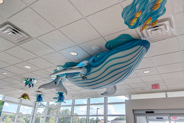 A large blue whale sculpture and other sea creatures are mounted on the ceiling of a bright room with white tiles and large windows.