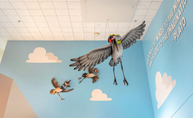 Three large, colorful bird sculptures fly against a blue wall with painted clouds and a Plato quote in white letters, creating a vibrant entrance for the undersea kids ministry. The ceiling is white with a grid pattern.