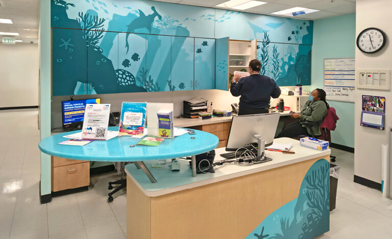 Colorful hospital reception area with two people, ocean-themed wall art reminiscent of an undersea kids ministry, computers, informational flyers, and a clock on the wall. One person is seated at the desk while another stands near the shelves.