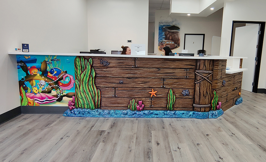 Reception desk with an underwater theme at a swim facility, featuring painted sea creatures, plants, and wood textures. Two people are partially visible behind the desk, and the flooring is gray wood-style laminate.