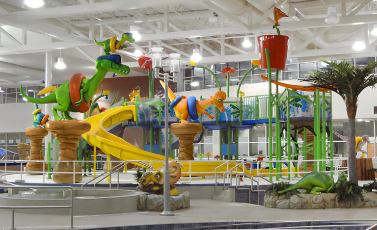 Indoor water play area with colorful dinosaur sculptures, slides, water sprayers, and a large tipping bucket under a high ceiling. Beach dental clinic decor elements blend seamlessly with palm tree decorations and splash zones throughout the space.