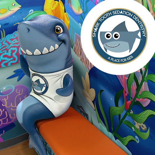 A friendly cartoon shark mascot sits on a bench in front of a colorful, pediatric dental decor-themed wall featuring the Shark Tooth Sedation Dentistry logo.