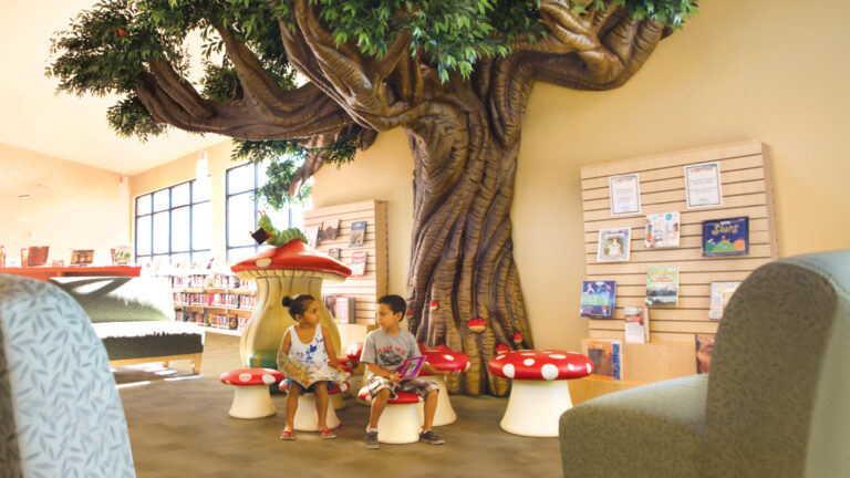 Two children sit on mushroom-shaped stools reading books beneath a large decorative tree painted as a wall mural inside a library.