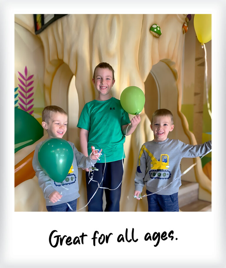 Three boys holding green balloons in front of a whimsical forest-themed wall mural play structure, with the text Great for all ages. written below them.