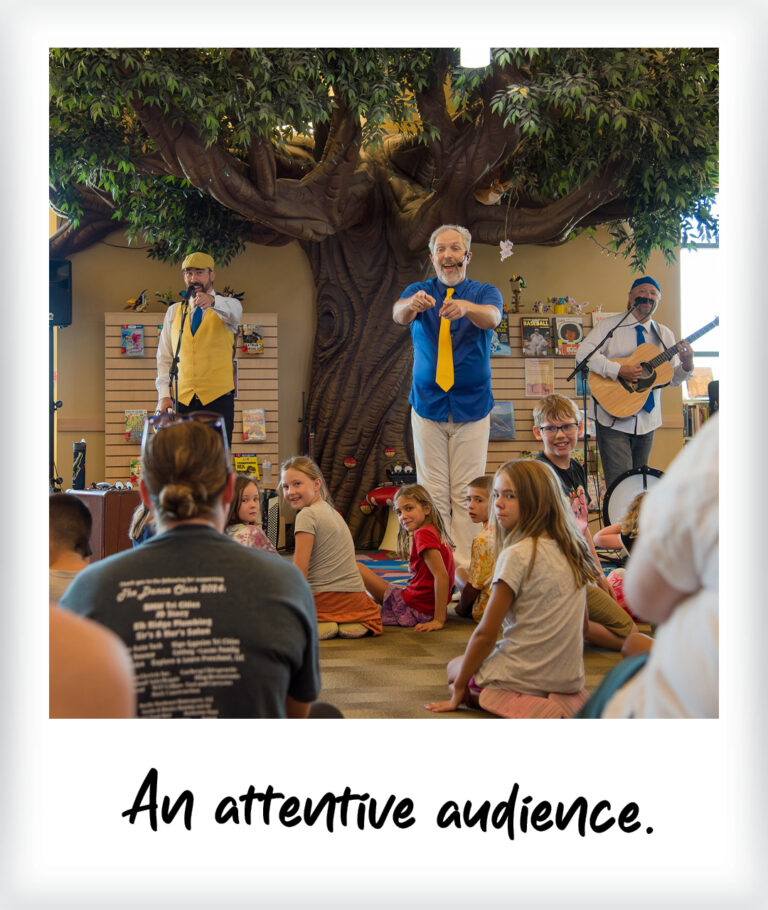 Three performers sing and play guitar for a group of children sitting on the floor in front of wall murals.